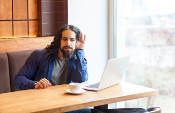 Man freelancer in a cafe having an online meeting struggling to hear holding his hand near his ear.