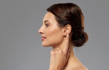 Profile of a woman with brown hair in a bun, touching her ear with her finger, against a gray background.