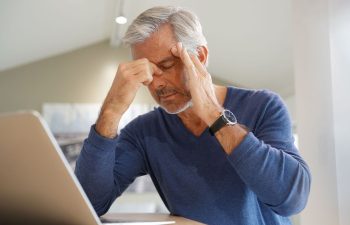 A fatigued mature man feeling unwell sitting at a table with laptop.